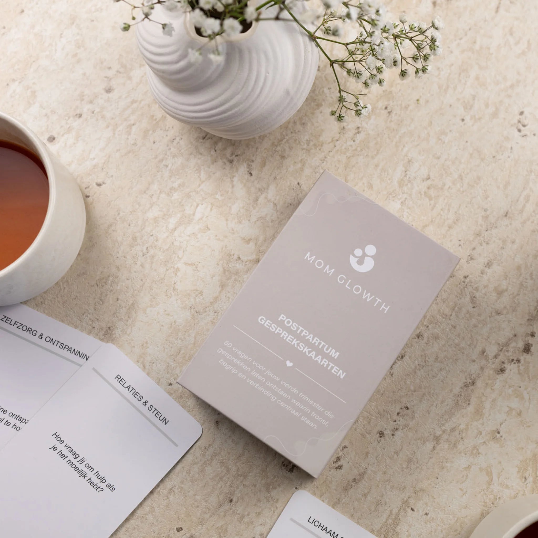 Postpartum gesprekskaarten van Mom Glowth uitgespreid naast bewaarbox, kopje thee en bloemen – cadeau voor nieuwe mama's in het vierde trimester